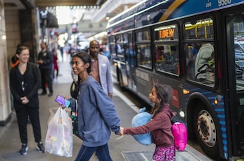 Una mujer y una niña caminan por una acera urbana. La mujer lleva un bolso transparente y la niña un peluche azul y una mochila rosa, junto a un autobús