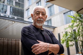 Hombre de mediana edad con cabello y barba blancos, camisa negra, brazos cruzados, mirando a cámara. De fondo, un edificio moderno y plantas verdes