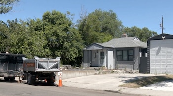 Vista exterior de una casa gris de un piso con techo a dos aguas y un garaje adyacente, con árboles verdes y un remolque de volteo en el camino de entrada