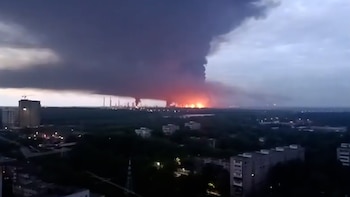 Incendio masivo en refinería industrial con llamas naranjas y humo negro que se eleva sobre un paisaje urbano al anochecer. Edificios visibles en primer plano