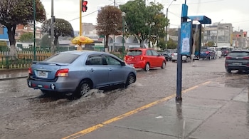 Automóviles que circulan sobre una calle cubierta de agua junto a una acera con semáforos, árboles y edificios al fondo.