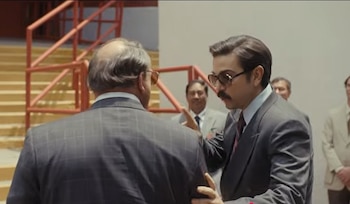 Primer plano de un hombre con bigote, gafas de sol y traje oscuro hablando con otro hombre de espaldas. Varios hombres en traje al fondo