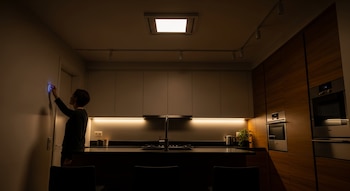 Una mujer de perfil de espaldas a una cocina moderna, ajusta un panel de control de iluminación azul brillante en la pared.