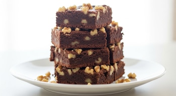 Pila de brownies húmedos de cacao y garbanzo, cortados en cuadrados, apilados sobre un plato blanco, con nueces esparcidas y un fondo luminoso.