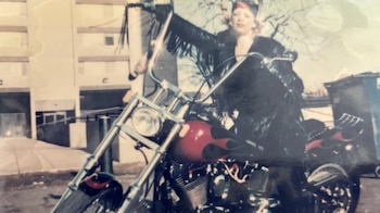 Mujer con vestimenta negra y sombrero en una motocicleta con tanque de combustible rojo con llamas negras, frente a un edificio y árboles