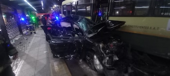 Un coche negro con daños severos en la parte delantera choca contra un autobús amarillo. Hay un bombero y personal de emergencia en el lugar