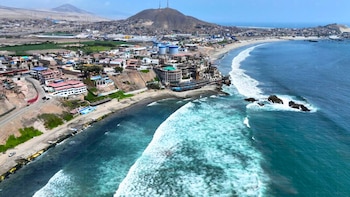 Vista aérea de la costa de Chancay con edificios urbanos a lo largo de la playa, olas rompiendo en el mar, rocas en la orilla y un cerro al fondo