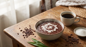 Un tazón de champorado humeante con crema, una taza de café, nibs de cacao, azúcar y hojas verdes sobre una mesa de madera rústica.