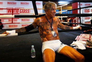 Mickey Rourke, de torso desnudo y tatuado, está sentado y sudoroso en un ring de boxeo, con un pantalón corto blanco y vendas en las manos