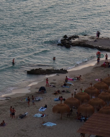 Vista aérea de playa con arena, personas bañándose en el mar, gente sentada y caminando, sombrillas de paja, y rocas en la orilla