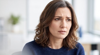 Una mujer de mediana edad con cabello castaño ondulado, vestida con un suéter azul oscuro, mira con el ceño fruncido en un entorno interior.