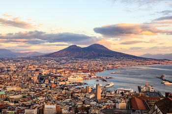 Vista aérea panorámica de la ciudad de Nápoles, su puerto con barcos, el mar, edificios y el Monte Vesubio bajo un cielo con nubes al atardecer