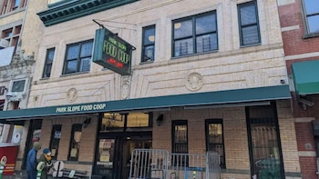 Fachada de un edificio de ladrillo claro con un toldo verde y un letrero luminoso que dice 'Park Slope Food Coop'. Hay ventanas oscuras y una entrada principal. Se ven barreras metálicas en la acera y dos personas cerca de la puerta