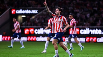 Futbolista con uniforme a rayas rojas y blancas que apunta al cielo sobre el césped, rodeado por otros jugadores y vallas publicitarias.