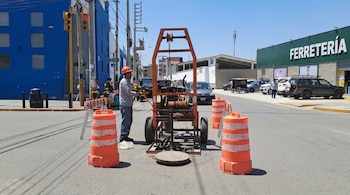 Un trabajador con casco opera maquinaria junto a un buzón abierto en la calle, rodeado por conos de seguridad color naranja