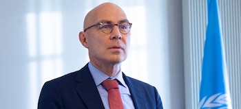 Primer plano de Volker Türk, un hombre calvo con gafas, traje azul marino y corbata roja, mirando hacia adelante. La bandera de la ONU se ve parcialmente a la derecha