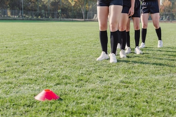 Primer plano de las piernas y pies de cuatro personas con shorts, medias y zapatillas deportivas negras y blancas en un campo de césped verde con un cono rojo