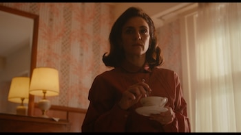 Una mujer de cabello oscuro y camisa rojiza sostiene una taza de té, mirando hacia arriba con expresión grave. Al fondo, papel tapiz y una lámpara