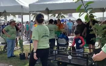 Varias personas con camisetas verdes atienden carpas blancas al aire libre junto a plantas en macetas y un carrito negro de metal