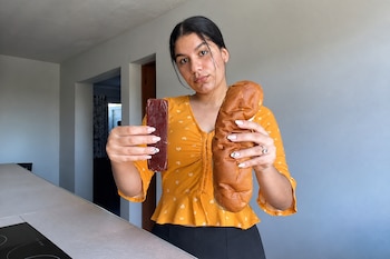 Una mujer viste una blusa naranja y sostiene un bloque rojizo rectangular en una mano y un pan alargado en la otra.