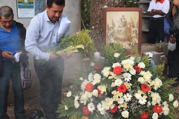 Dos hombres de pie; uno sostiene un ramo de flores y otro un folleto. Un cuadro con una imagen religiosa y arreglos florales con velas