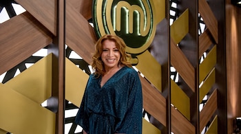 Una mujer de cabello castaño y vestido azul posa sonriente frente al logotipo de MasterChef y paredes decoradas de madera.