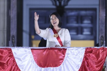 Una mujer de pelo oscuro con vestido claro y banda presidencial roja y blanca sonríe y saluda desde un balcón con barandilla cubierta de tela roja y blanca