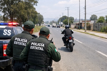 Dos policías estatales de espalda con chalecos y gorras, junto a una camioneta policial, observan a un motociclista que se aleja por una carretera.