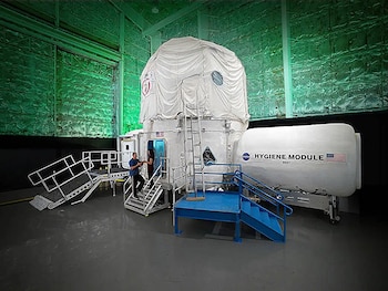 Módulo de hábitat inflable blanco y módulo de higiene cilíndrico, con dos personas en las escaleras de acceso, dentro de una gran instalación de la NASA