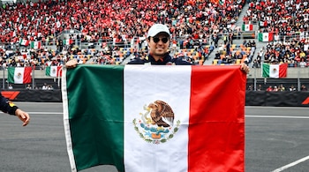 Sergio Pérez con gorra y gafas de sol sostiene una bandera mexicana extendida frente a gradas repletas de espectadores.