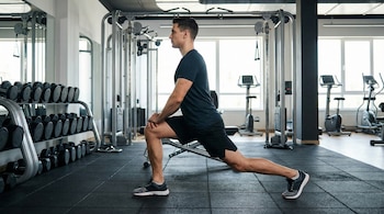 Um jovem de perfil está fazendo estocadas em uma academia, com as mãos nos joelhos dianteiros, cercado por pesos e máquinas.