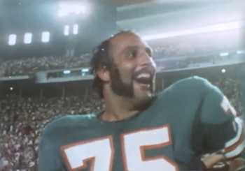 Primer plano de Manny Fernandez, un hombre barbudo con camiseta verde de fútbol americano número 75, sonriendo ampliamente en un estadio