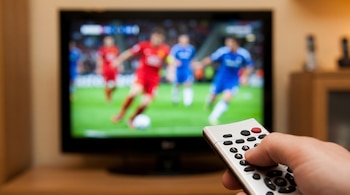 Mano sosteniendo un control remoto blanco con botones negros y rojos en primer plano; al fondo, un televisor desenfocado muestra un partido de fútbol.