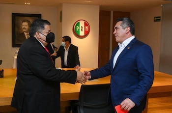 Dos hombres se estrechan las manos en una sala de reuniones. Uno lleva mascarilla y traje oscuro, el otro traje azul. El logo del PRI y un cuadro están en la pared