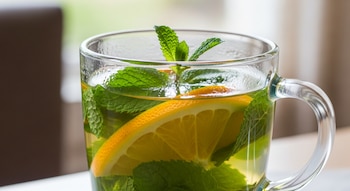 Primer plano de una taza transparente de té verde, con hojas de hierbabuena fresca y una rodaja de naranja flotando, bajo luz natural.