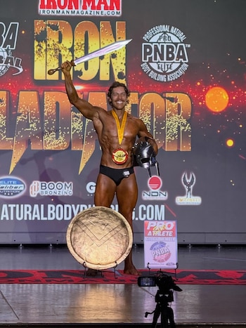 Joseph Baena, fisicoculturista, posa en el escenario con una medalla, sosteniendo una espada en alto y un casco de gladiador en la otra mano