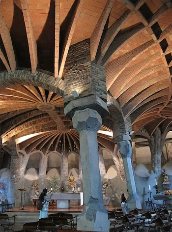 Interior de la Cripta de la Colonia Güell con arcos de ladrillo, columnas inclinadas de piedra, bancos de madera y algunas personas admirando la arquitectura