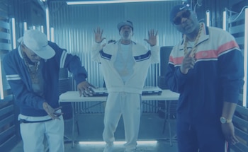 Tres hombres con gorras, gafas de sol y cadenas de oro posan en un estudio con luces azules. Uno manipula una mesa de mezclas, otro levanta las manos, el tercero señala