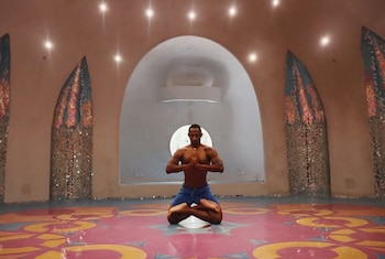Hombre practicando yoga en un tapete azul junto a un río verde. Viste camiseta beige y shorts azules. Al fondo, edificios coloridos y montañas