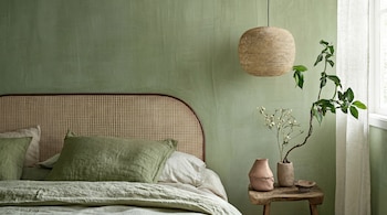 Dormitorio con pared verde, cama con cabecero de ratán, ropa de cama verde y beige, lámpara colgante de fibra natural y una mesa de madera con plantas.