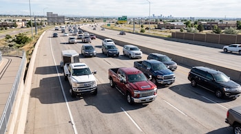 Camionetas y automóviles avanzan por los carriles de una autopista con la silueta de una ciudad en el horizonte.