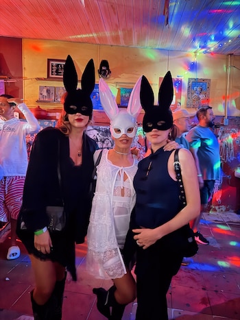 Tres mujeres con máscaras de orejas de conejo, dos de color negro y una blanca en el centro, posan en un recinto iluminado con luces de colores