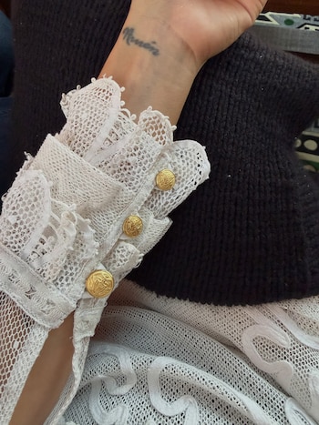Un antebrazo con un tatuaje en la muñeca lleva una manga de encaje blanco con tres botones dorados sobre una superficie de tejido negro