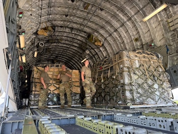 Tres personas con uniforme militar dentro de un avión de carga junto a grandes paletas de suministros envueltos y asegurados con redes