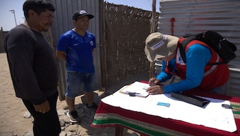 Tres hombres al aire libre: uno de Cofopri escribe en un portapapeles sobre una mesa, mientras dos hombres observan, con fondo de cercas metálicas y de caña