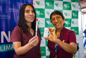 Dos mujeres sostienen un hisopo de prueba médica y un frasco blanco. Detrás, banners con logotipos de MINSA DIRIS LIMA SUR y Línea 1 Metro de Lima