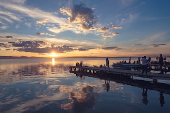 Vista de un lago al atardecer con el sol en el horizonte, nubes en el cielo y su reflejo en el agua. Un muelle de madera con personas y embarcaciones
