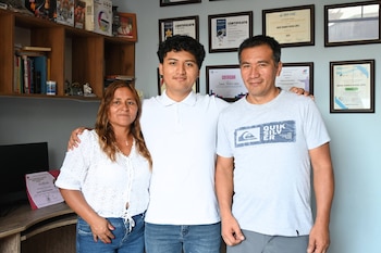 David Rivera (centro), un joven sonriente de 19 años, posa con su madre (izquierda) y su padre (derecha). Detrás, una pared con diplomas y estantes