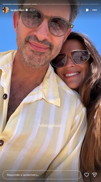 Primer plano de un hombre con barba y gafas de sol, con una mujer sonriente detrás apoyada en su hombro, ambos al aire libre con cielo azul
