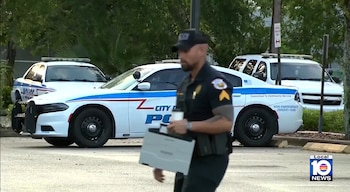 Oficial de policía con uniforme oscuro camina con un objeto plateado en sus manos; detrás, dos vehículos policiales blancos con logotipos azules y rojos